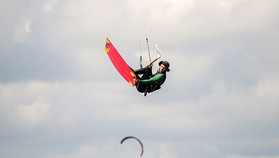 kitesurfer rides the waves at his home spot in the bay. An athlete in a panama hat performs a trick in the air while flying up against the background of a cloudy sky